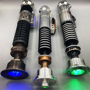 May include: Three lightsaber replicas with different colored lights. The lightsabers are silver and black with copper accents. The lightsabers are lit up with blue, green, and green lights.