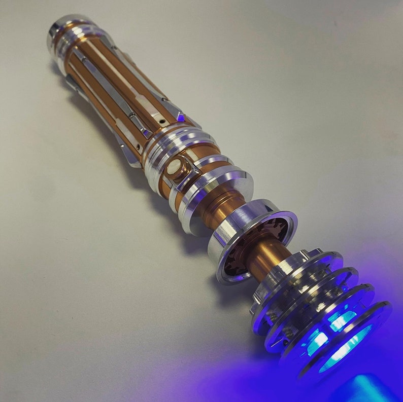 May include: A lightsaber with a gold and silver body and a blue light saber blade. The lightsaber is disassembled and the blade is lit.