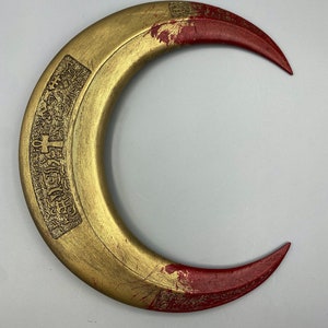 May include: A crescent-shaped, golden-colored object with red tips. The object has an engraved design on the inner curve. The surface shows signs of wear, with some paint chipping. The overall design suggests a decorative or ceremonial item.