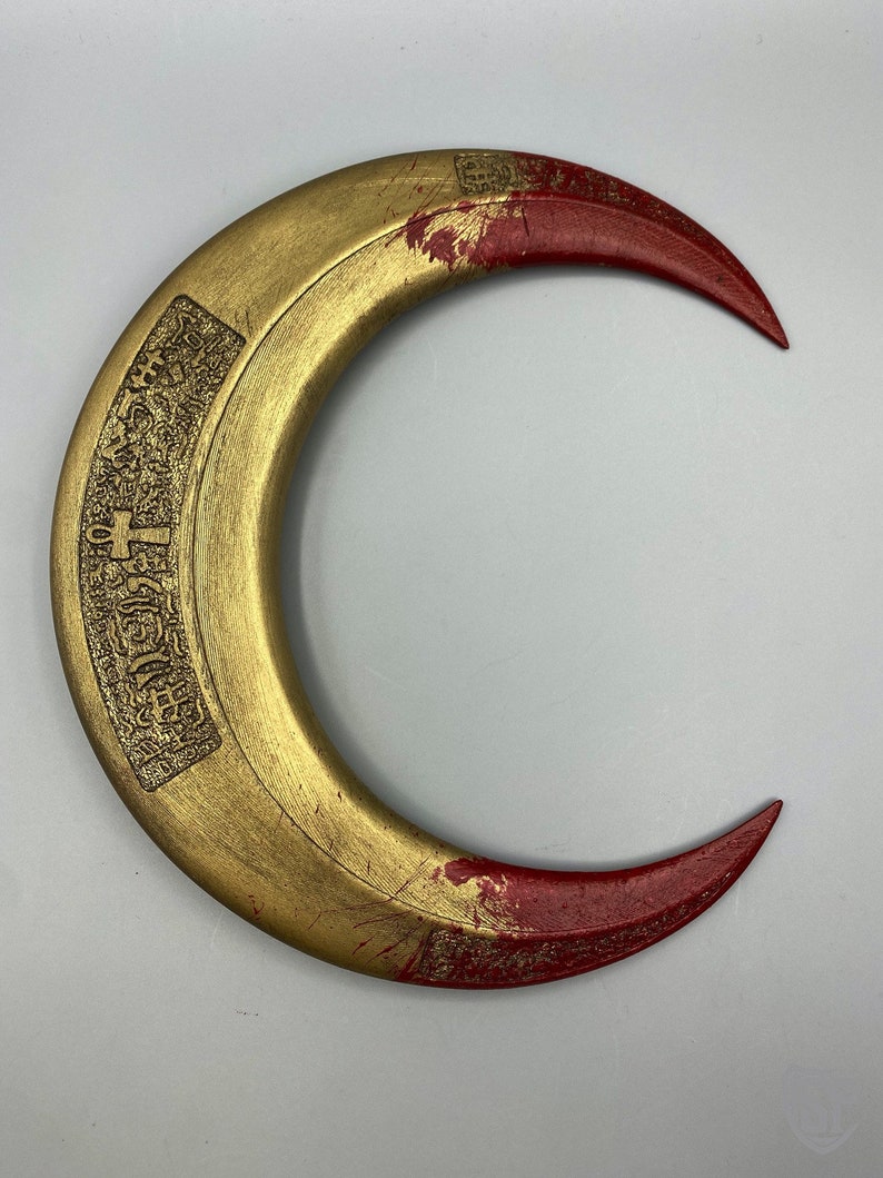 May include: A crescent-shaped, golden-colored object with red tips. The object has an engraved design on the inner curve. The surface shows signs of wear, with some paint chipping. The overall design suggests a decorative or ceremonial item.