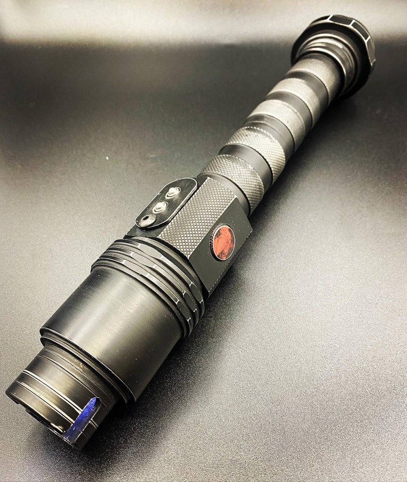 May include: A black lightsaber prop with a red button and a textured grip. The lightsaber is made of metal and has a black and silver finish.