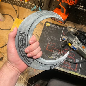 May include: A crescent-shaped, gray prop weapon with etched details and a handle. The prop has a metallic appearance and is held in a person's hand. The weapon has a sharp, curved blade and is likely used for cosplay or display.