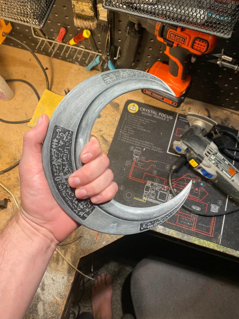 May include: A crescent-shaped, gray prop weapon with etched details and a handle. The prop has a metallic appearance and is held in a person's hand. The weapon has a sharp, curved blade and is likely used for cosplay or display.
