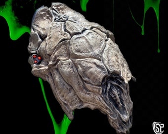 May include: A detailed, gray, textured, alien creature head with red eyes. The creature's head is against a black background with green slime dripping down.