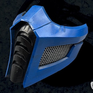 Puede incluir: Una máscara azul y negra con un diseño geométrico. La máscara presenta un marco azul, una malla de panal y una sección inferior negra texturizada. Probablemente para cosplay o disfraces.
