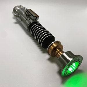 May include: A silver and black lightsaber with a green light saber blade. The lightsaber has a gold metal piece at the end of the handle.