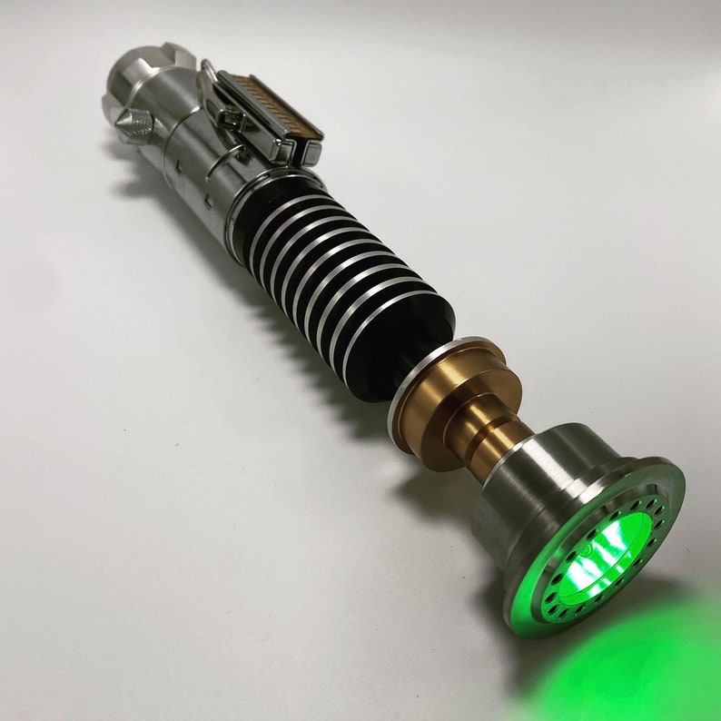 May include: A silver and black lightsaber with a green light saber blade. The lightsaber has a gold metal piece at the end of the handle.