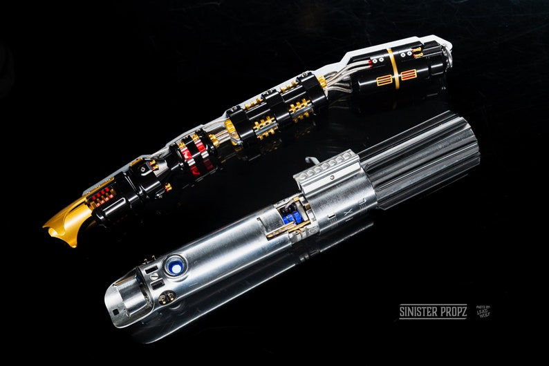 May include: Two disassembled lightsabers, one with a gold and black hilt and the other with a silver hilt. The lightsabers are on a black surface. The text "SINISTER PROPZ" is visible in the lower right corner of the image.