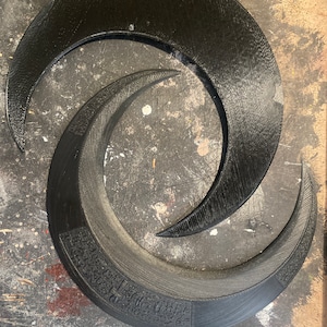 May include: Two black, crescent-shaped objects, one larger than the other, are arranged in an overlapping spiral. The larger piece has a textured surface and a decorative band with etched details. The smaller piece is smooth.