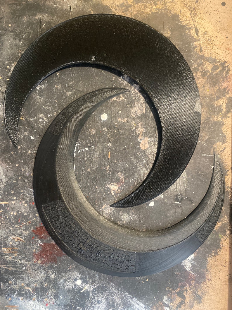 May include: Two black, crescent-shaped objects, one larger than the other, are arranged in an overlapping spiral. The larger piece has a textured surface and a decorative band with etched details. The smaller piece is smooth.
