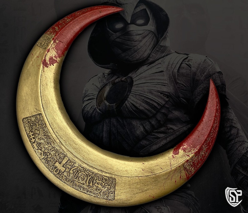 May include: A gold crescent-shaped weapon with red tips, possibly a prop from a movie or comic book. The weapon has engraved details and is set against a dark background with a character in the background.