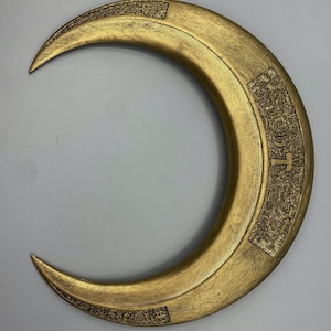 May include: A gold-colored crescent-shaped prop with etched details. The prop has a textured surface and appears to be made of metal or a similar material. The design includes rectangular panels with intricate patterns and symbols.