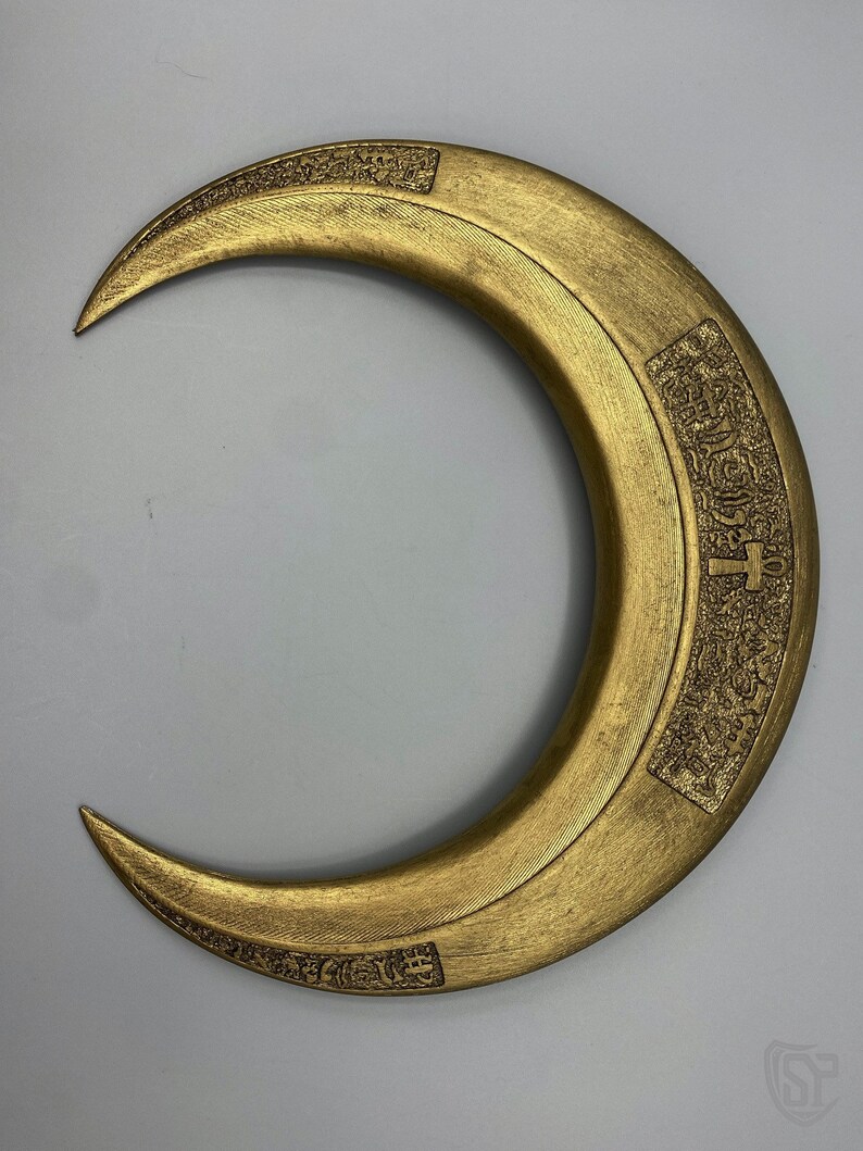 May include: A gold-colored crescent-shaped prop with etched details. The prop has a textured surface and appears to be made of metal or a similar material. The design includes rectangular panels with intricate patterns and symbols.