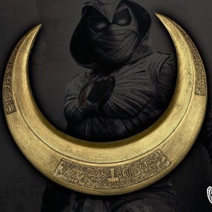 May include: A gold-colored crescent-shaped weapon with intricate engraved designs. The weapon is set against a dark background with a character in a hooded costume. The crescent has a flat, wide profile and appears to be made of metal.