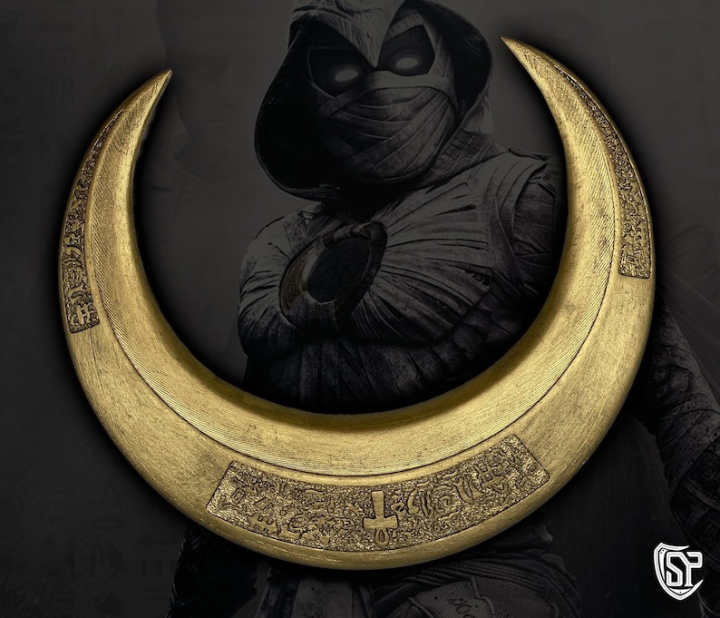 May include: A gold-colored crescent-shaped weapon with intricate engraved designs. The weapon is set against a dark background with a character in a hooded costume. The crescent has a flat, wide profile and appears to be made of metal.