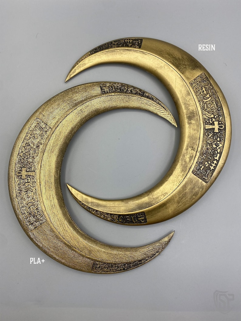 May include: Two golden crescent-shaped objects with intricate engraved patterns. The top object is labeled "RESIN" and the bottom object is labeled "PLA+". The objects are displayed on a gray background.