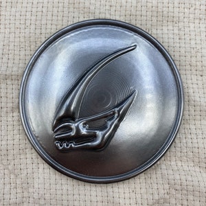 May include: A circular, metallic gray emblem with a raised, stylized design. The design features a sharp, curved element and a detailed, skeletal-like structure. The emblem has a brushed metal finish and is set against a textured, beige background.