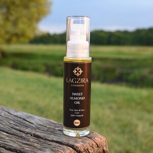 Op de afbeelding: Een doorzichtige glazen fles met een zwart etiket waarop "LAGZIRA LONDON SWEET ALMOND OIL Hair, Skin & Face Care 100% Natural 50ml" staat. De fles staat op een houten oppervlak.