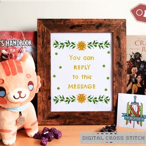 Op de afbeelding: Een digitaal kruissteekpatroon met een bericht dat luidt "You can REPLY to this MESSAGE". Het patroon is omgeven door groene bladeren en gele bloemen. Het patroon wordt getoond in een houten frame en wordt vergezeld door een pluche kattenknuffel, een Dungeons and Dragons Player's Handbook en een Critical Role boek.