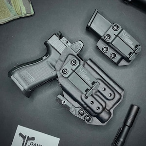 May include: Black handgun with a red dot sight, a black holster, and a magazine holder. A black flashlight and a patch with the text "Davis Tactical" are also visible. The items are on a dark surface.