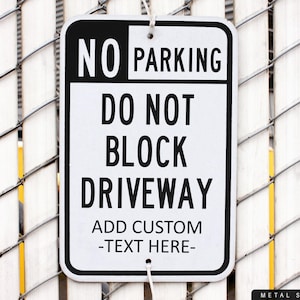 May include: A rectangular black and white metal sign with rounded corners, hanging on a chain-link fence. The sign reads "NO PARKING DO NOT BLOCK DRIVEWAY" with space to add custom text.