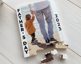 May include: A white and brown photo puzzle with a picture of a father and child on a beach. The puzzle is made up of individual square pieces. The text "Father's Day 2023" is printed on the side of the puzzle.