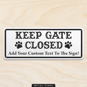 May include: A rectangular metal sign with a white background and black border. The sign reads "KEEP GATE CLOSED" with paw prints and "Add Your Custom Text To The Sign!" in black text. The sign is mounted on a light wood surface.