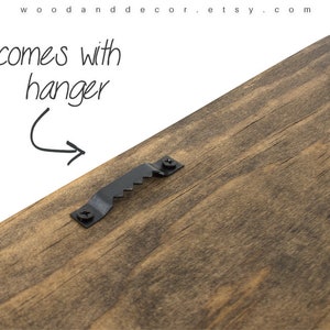 May include: The back of a wooden sign with a black metal hanger attached. The sign is made of dark brown wood with a natural grain pattern.