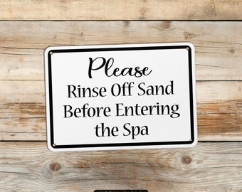 May include: A rectangular white metal sign with a black border and the words "Please Rinse Off Sand Before Entering the Spa" in black lettering. The sign is mounted on a wooden background.