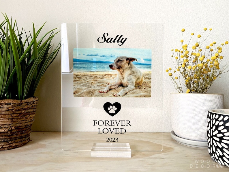 Peut inclure: Un cadre photo en acrylique transparent avec un c&oelig;ur noir en forme de patte et le texte "Forever Loved 2023". Le cadre contient une photo d'un chien nomm&eacute; Sally sur une plage.