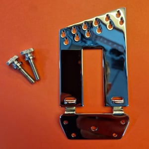 May include: A shiny chrome-plated guitar bridge with a rectangular opening and multiple mounting holes. Two silver screws are positioned to the left of the bridge. The background is a solid orange color.