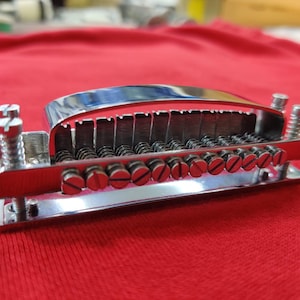 May include: A chrome-plated guitar bridge with adjustable saddles and string slots. The bridge has a curved top and is mounted on a red surface. The bridge is designed to hold the strings in place and allow for adjustments to the instrument's intonation.