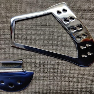 May include: Two shiny chrome metal parts. One is a rectangular frame with a curved handle and several small holes. The other is a smaller, curved piece with three holes. Both pieces reflect light.