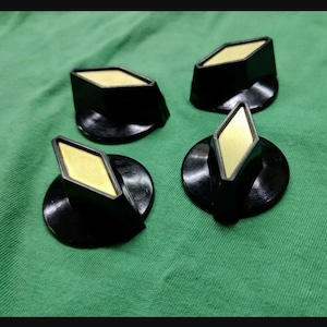 May include: A set of four black control knobs with a circular base and a diamond-shaped top. Each knob features a flat, gold-colored inlay centered on the diamond-shaped face. The knobs have a smooth, glossy finish and a tapered, angular design.