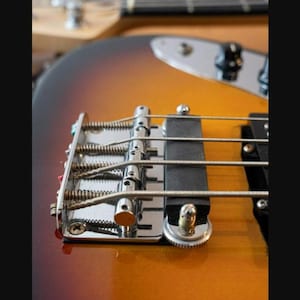 Könnte beinhalten: Eine Sunburst-E-Bass-Gitarrenbrücke mit vier Metallsätteln inklusive Spiralfedern, einer schwarzen Tonabnehmerabdeckung und silbernen Beschlägen. Der Instrumentenkorpus zeigt einen Farbverlauf von Dunkelbraun zu Bernsteingelb.
