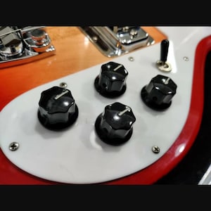 May include: A close-up view of an electric guitar body featuring a white pickguard, four black control knobs with silver indicator lines, a small black toggle switch, and a chrome bridge assembly set against a sunburst wood finish.