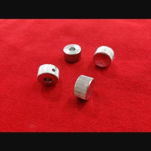 Peut inclure: Quatre petits boutons cylindriques en métal argenté avec un bord texturé et moleté. Chaque bouton présente une surface supérieure plate et une ouverture circulaire centrale, l'un d'eux montrant un petit trou latéral pour une vis.