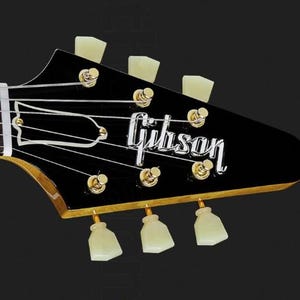 May include: Close-up of the headstock of a black electric guitar with gold hardware. The guitar has six tuning pegs with white plastic buttons. The Gibson logo is visible on the headstock.