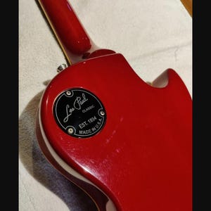 Pode incluir: Um corpo de guitarra elétrica vermelho brilhante com uma tampa circular preta para a cavidade de controlo. A tampa apresenta texto branco que diz Les Paul CLASSIC, EST. 1894 e MADE IN U.S.A. O braço da guitarra estende-se para cima.