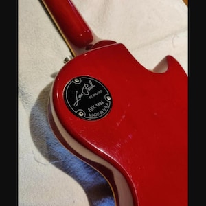 May include: A glossy cherry red electric guitar body featuring a circular black control cavity cover plate. The plate is printed with the text Les Paul Standard, EST. 1894, and MADE IN U.S.A. in white lettering, secured by four small silver screws.