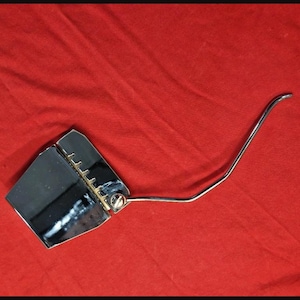 Può includere: Un sistema di ponte tremolo per chitarra cromato caratterizzato da una piastra di base rettangolare con fessure per le corde e un lungo braccio a leva in metallo curvo attaccato. La superficie metallica è altamente riflettente.