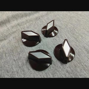 May include: Four dark brown control knobs with a circular base and a diamond-shaped top. Each knob features a flat, silver-colored metallic inlay centered on the diamond-shaped handle, providing a contrast to the dark brown material of the knob.