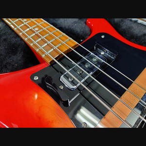 Puede incluir: Un bajo eléctrico de color rojo brillante con acabado lustroso, golpeador negro y una pastilla cromada. El instrumento cuenta con un diapasón de madera clara con trastes metálicos visibles y cuatro cuerdas plateadas sobre el cuerpo.