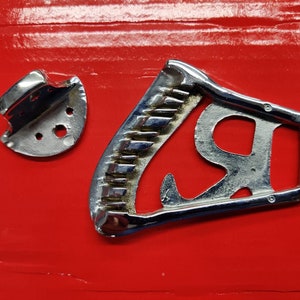 May include: Two silver metal objects on a red background. One object is a small, round, metal piece with a hole in the center. The other object is a larger, curved, metal piece with a serrated edge and a hole in the center.