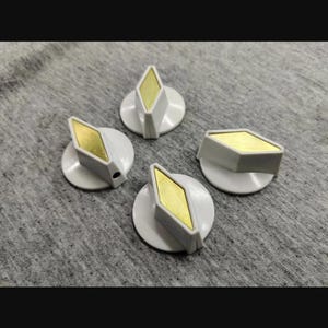 May include: Four white control knobs with a diamond-shaped gold-colored metal inlay on the top surface. Each knob features a circular base and a raised, angled center section designed for manual adjustment, showcasing a clean and functional design.