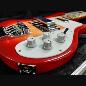 May include: A fireglo red electric bass guitar featuring a white pickguard, three white control knobs, a chrome bridge cover, and a black toggle switch. The instrument has a glossy finish, a metal output jack plate, and visible metal strings.
