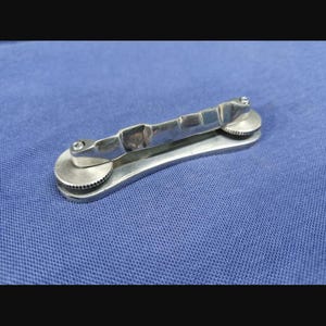 Op de afbeelding: Een zilverkleurige metalen sleutelorganizer met een slank, langwerpig lichaam en twee ronde, getextureerde knoppen aan elk uiteinde. Het apparaat heeft een centrale brugstructuur die de twee uiteinden verbindt om sleutels stevig vast te houden.