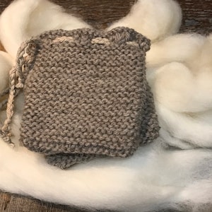 May include: A small, gray knitted drawstring bag with a white drawstring. The bag is sitting on a pile of white wool.