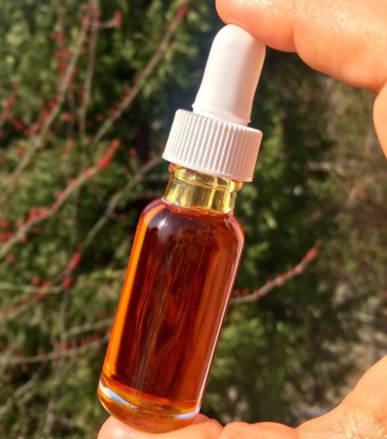 May include: A small amber glass bottle with a white dropper top, filled with a dark orange liquid. The bottle is held in a hand against a blurred green background.