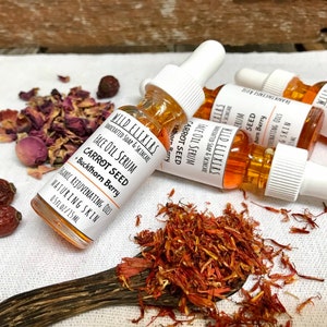May include: A close-up of a small glass bottle with a dropper filled with a golden liquid. The bottle is labeled "Wild Elixirs Face Oil Serum Carrot Seed + Buckthorn Berry". The bottle is surrounded by dried rose petals and saffron.